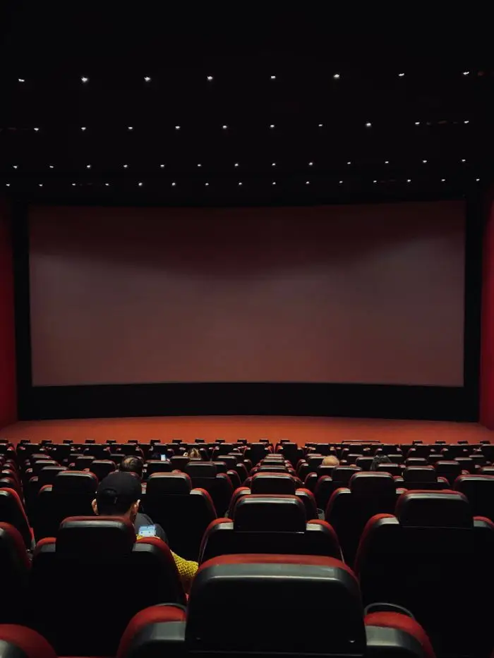Dimly lit cinema hall with red seats and a blank screen. Minimal audience.